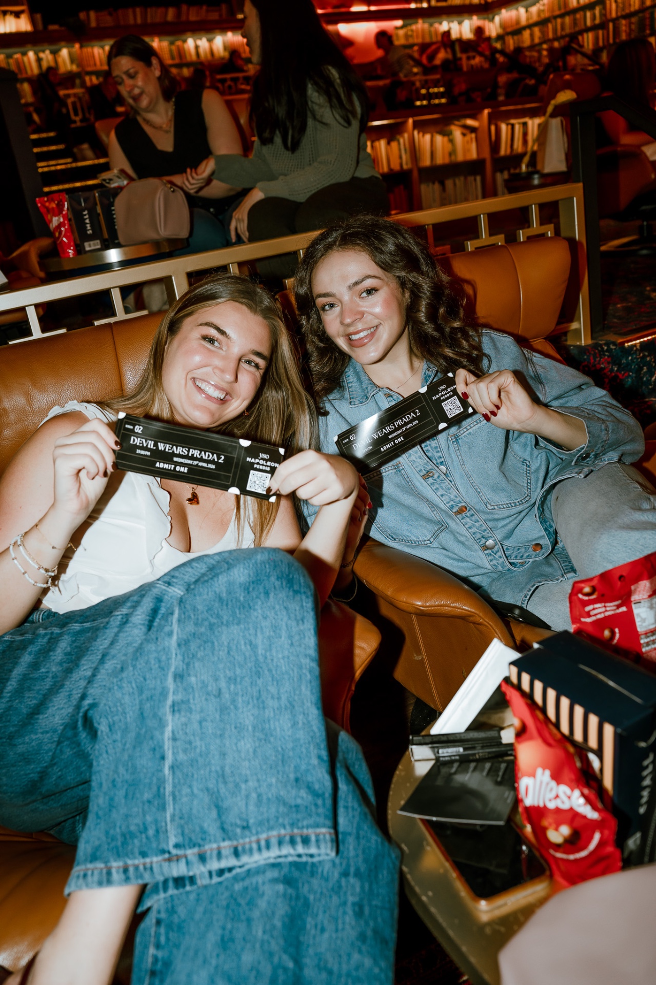 Two friends hold Devil Wears Prada 2 admit-one tickets at the YSO cinema club premiere night in a gold class library-style lounge in Sydney, surrounded by Maltesers and Napoleon Perdis goodie bags.