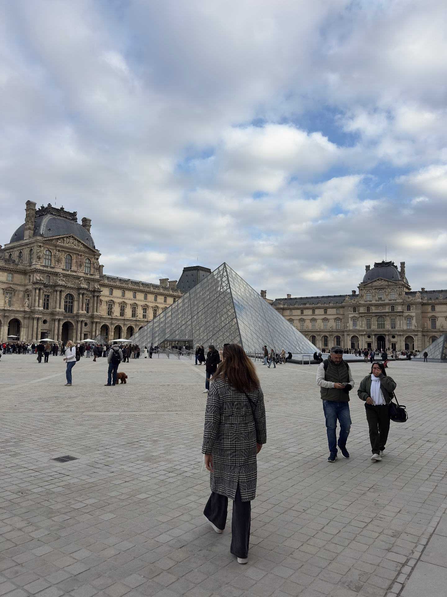 A woman in a houndstooth coat walks across the Louvre courtyard towards the glass pyramid with the historic palace wings behind her.