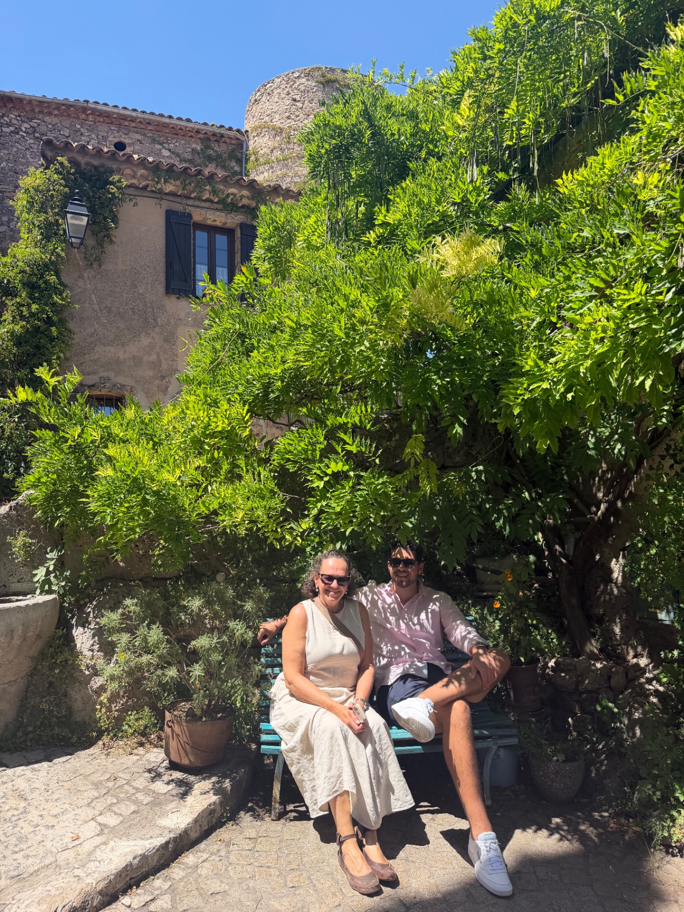 Two travellers sitting on a small turquoise bench beneath a cascade of wisteria in Tourtour, a medieval hilltop village in Provence, with a stone tower of the old village rising behind.