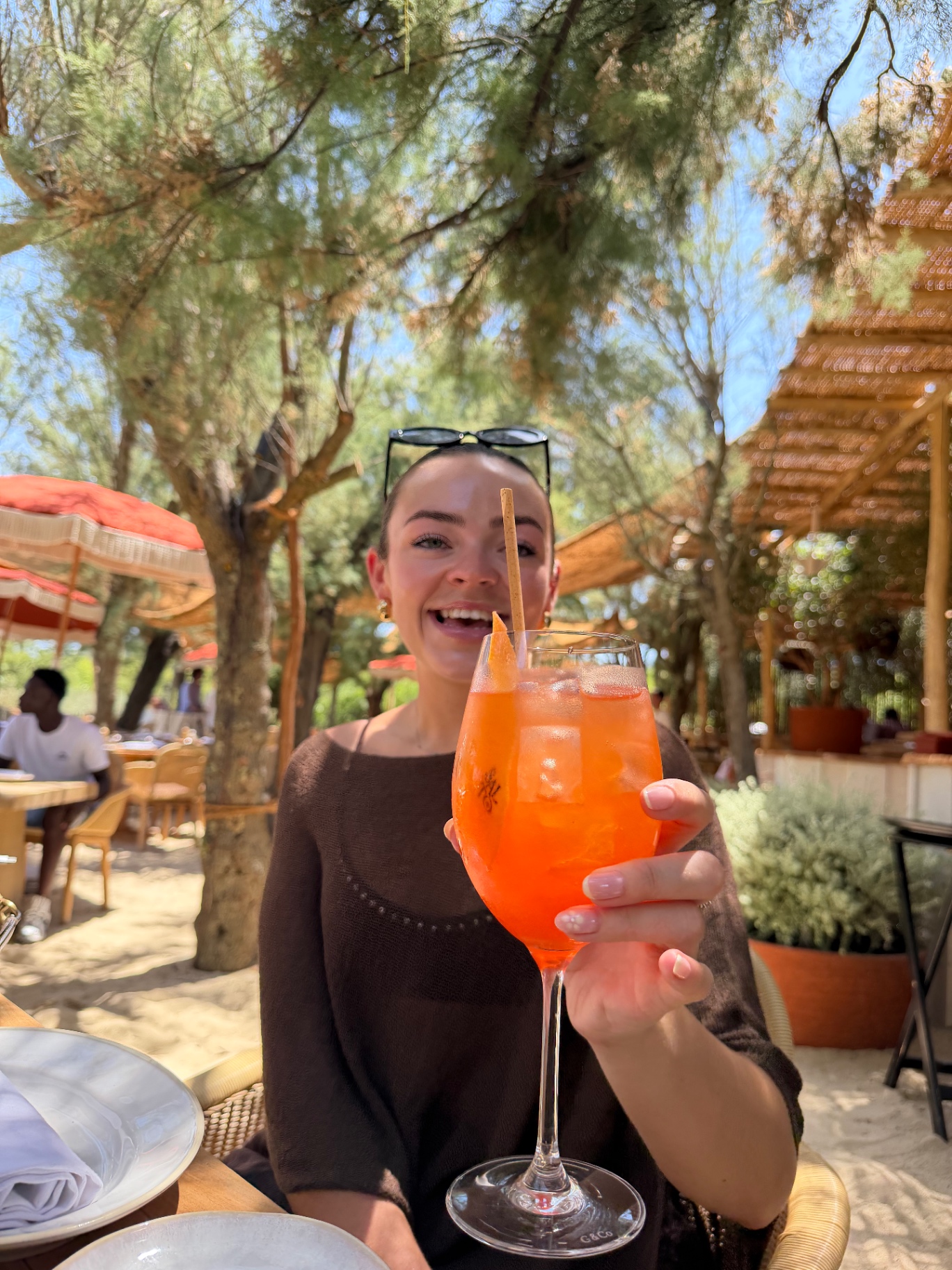 Holly holding SALTO's signature burnt-pineapple spritz at the beach club restaurant, with tamarisk trees and terracotta parasols in the background.