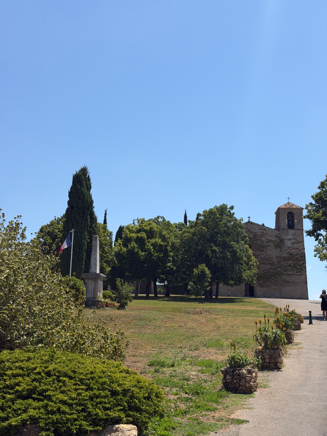 The exterior of Saint-Denis church on the hill at Tourtour, with cypress trees, a French tricolore flag and a stone war memorial in the foreground.