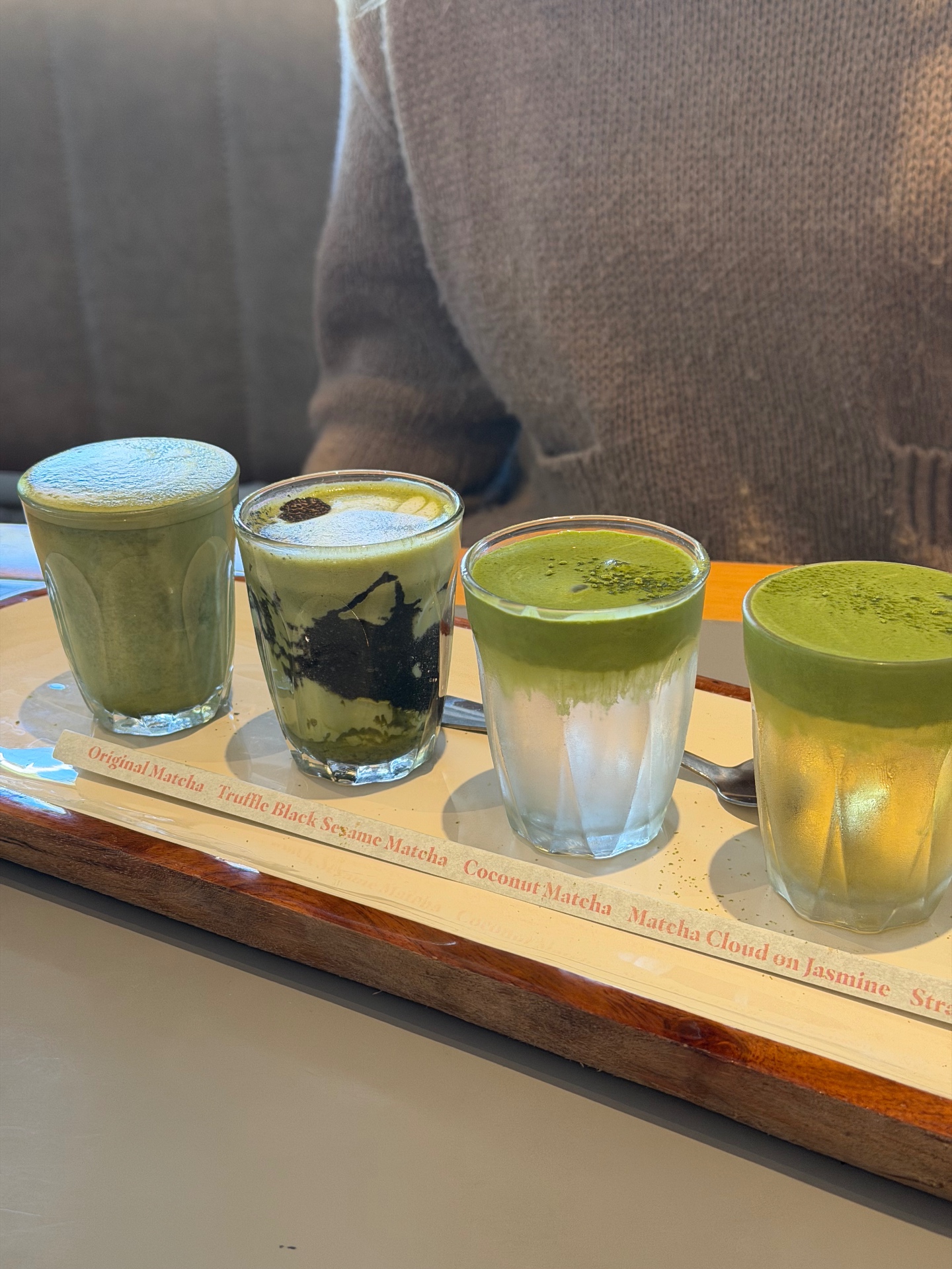 The winter matcha flight at The Wyatt in Gordon — Original Matcha, Truffle Black Sesame Matcha, Coconut Matcha and Matcha Cloud on Jasmine in piccolo glasses on a wooden tray with a printed label strip.