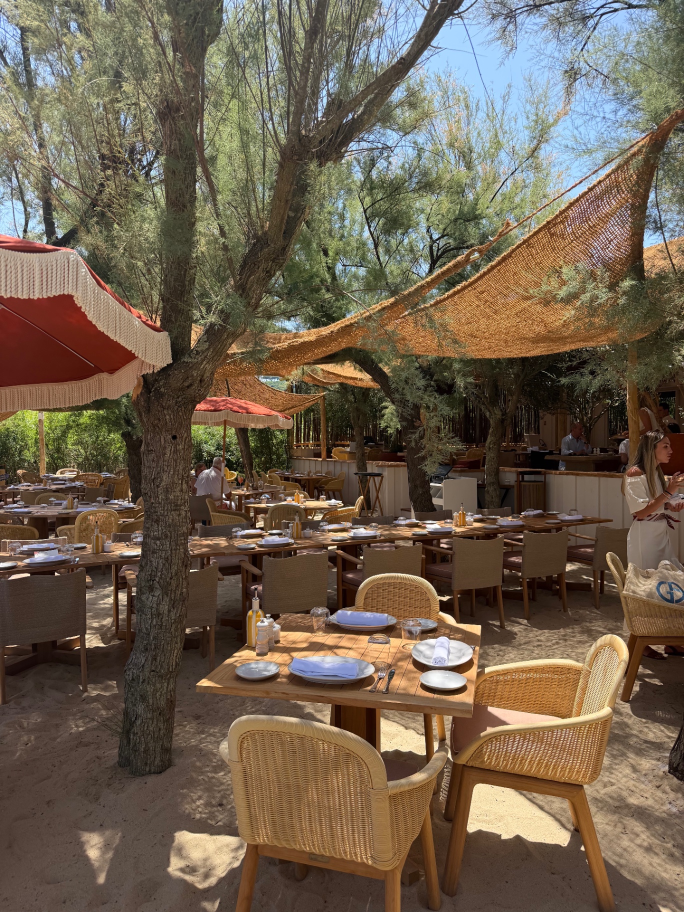 Tables set for lunch on the sand at SALTO, scattered between tamarisk trees with sail-like woven shades and terracotta parasols overhead.