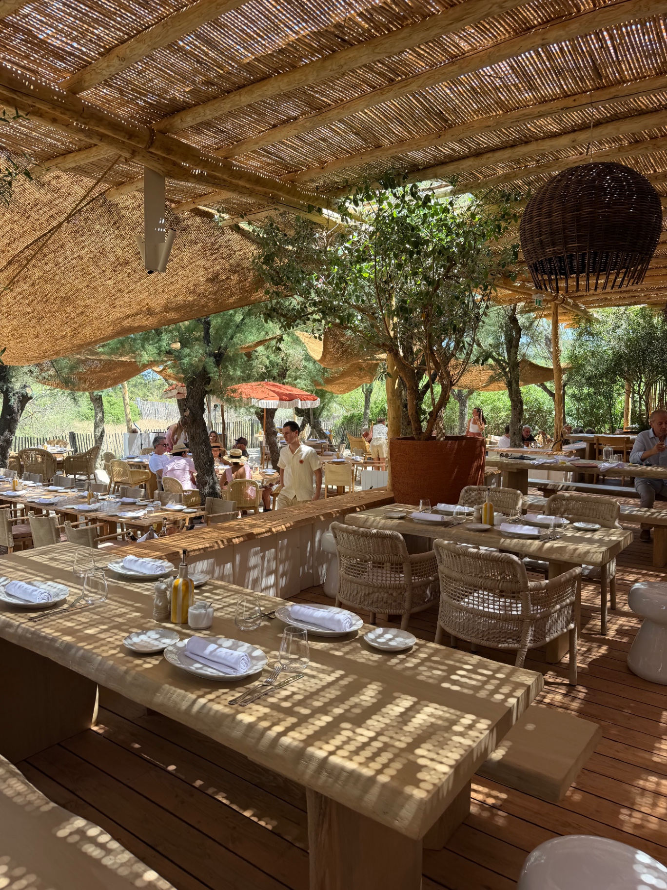 The covered restaurant deck at SALTO beach club with long timber tables, woven rattan chairs and a bamboo ceiling, dappled with sunlight.