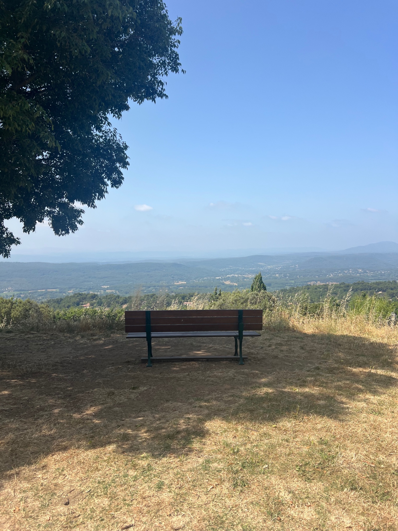 A wooden bench under a tree on the ridge at Tourtour, looking out across the Var valley and a hazy line of distant hills in Provence.