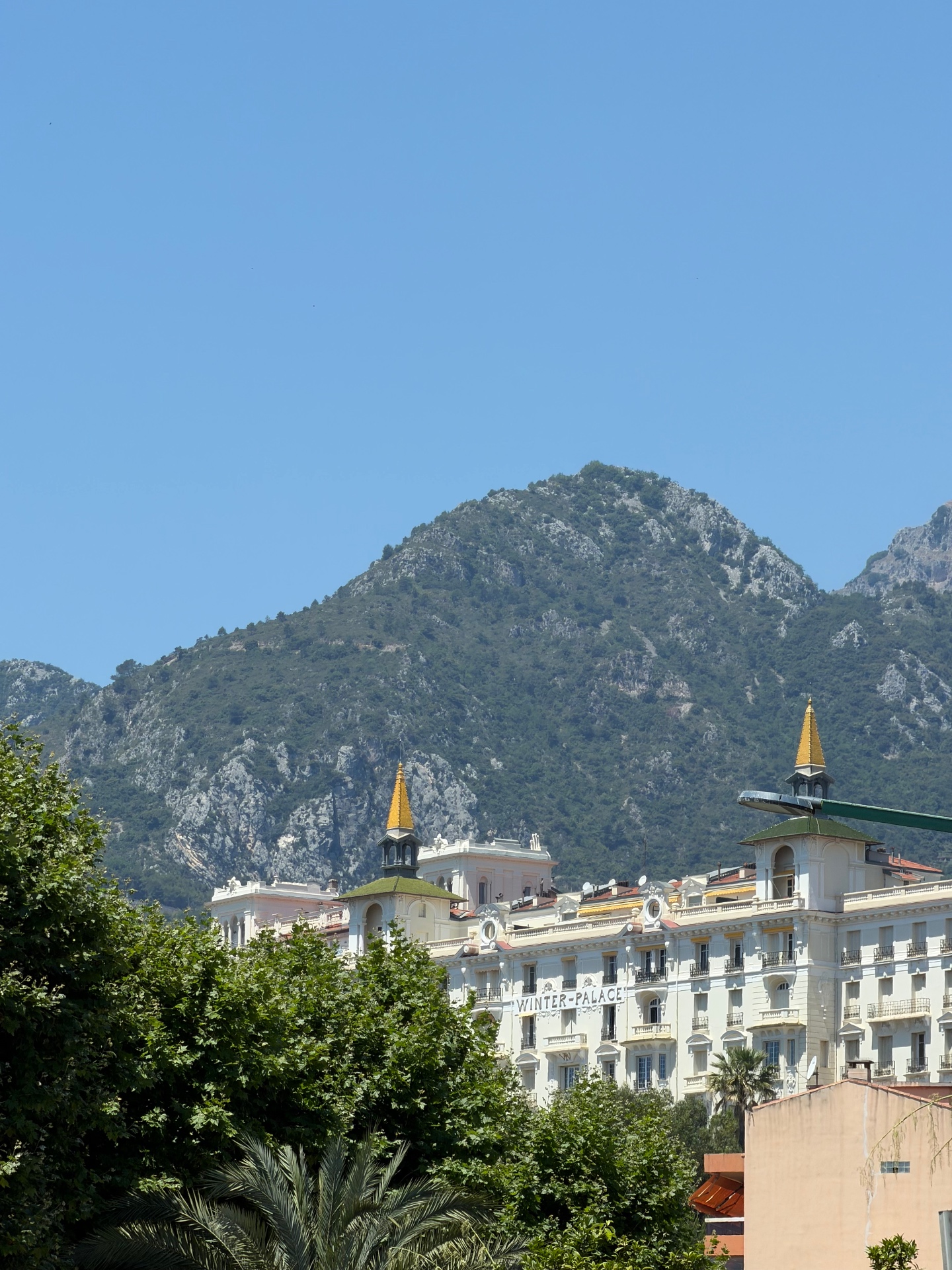 The white Belle Époque façade of the Winter Palace in Menton with its two gold-spired turrets set against a steep, rocky mountain backdrop.