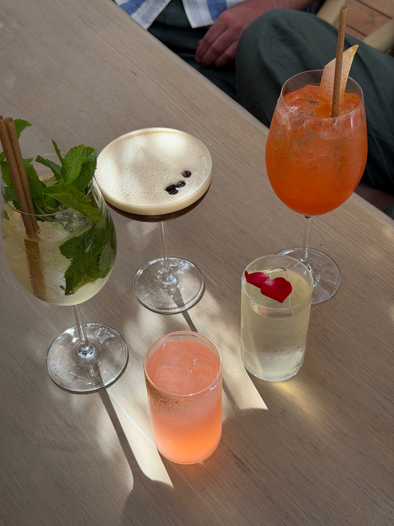 A spread of cocktails on a wooden table at SALTO including a mojito, espresso martini, Aperol spritz, rose-petal highball and a paloma in afternoon sunlight.