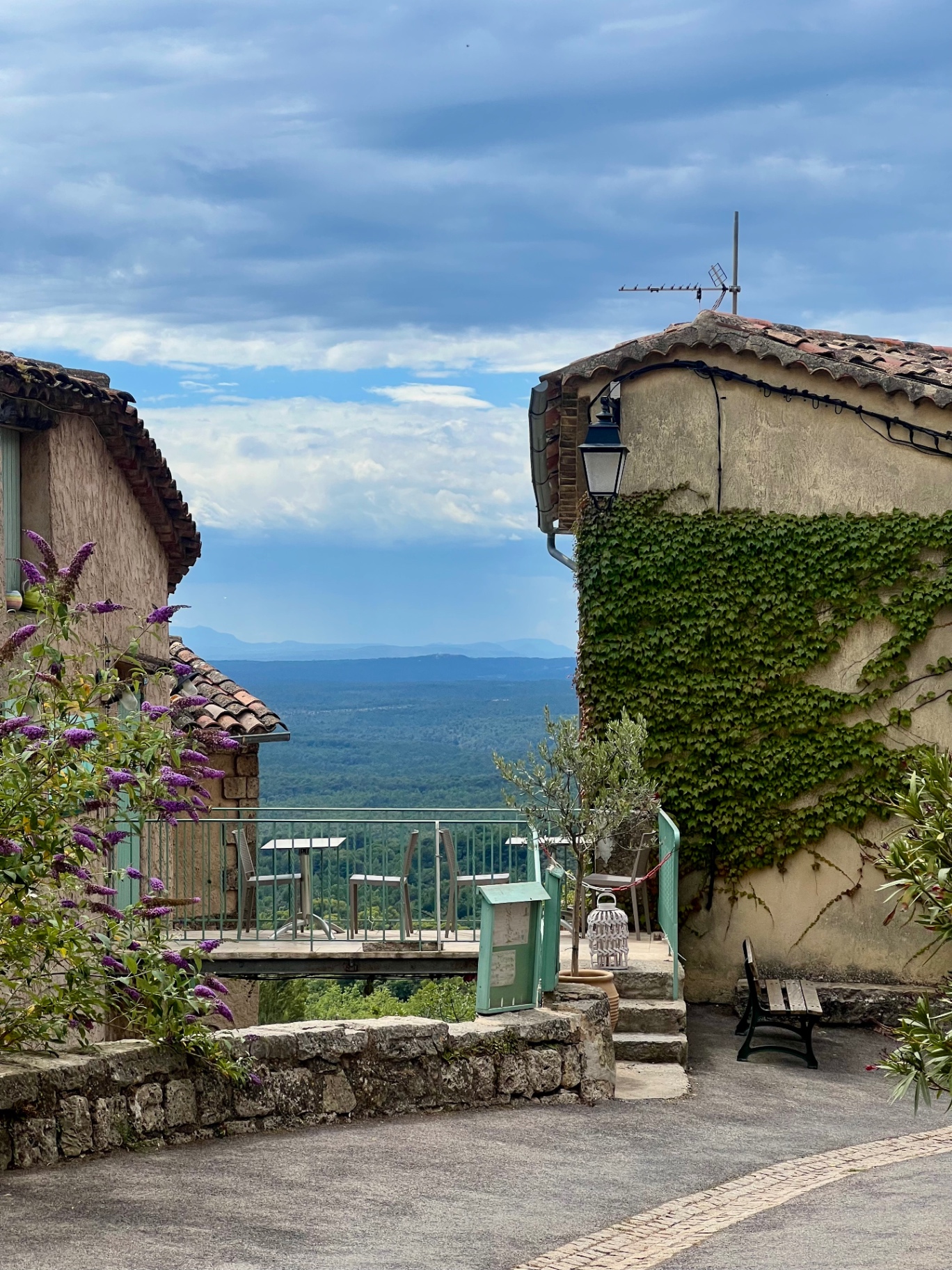 A small terrace café in Tourtour overlooking the green Var valley below, framed by two ivy-covered Provençal stone houses with terracotta roofs.