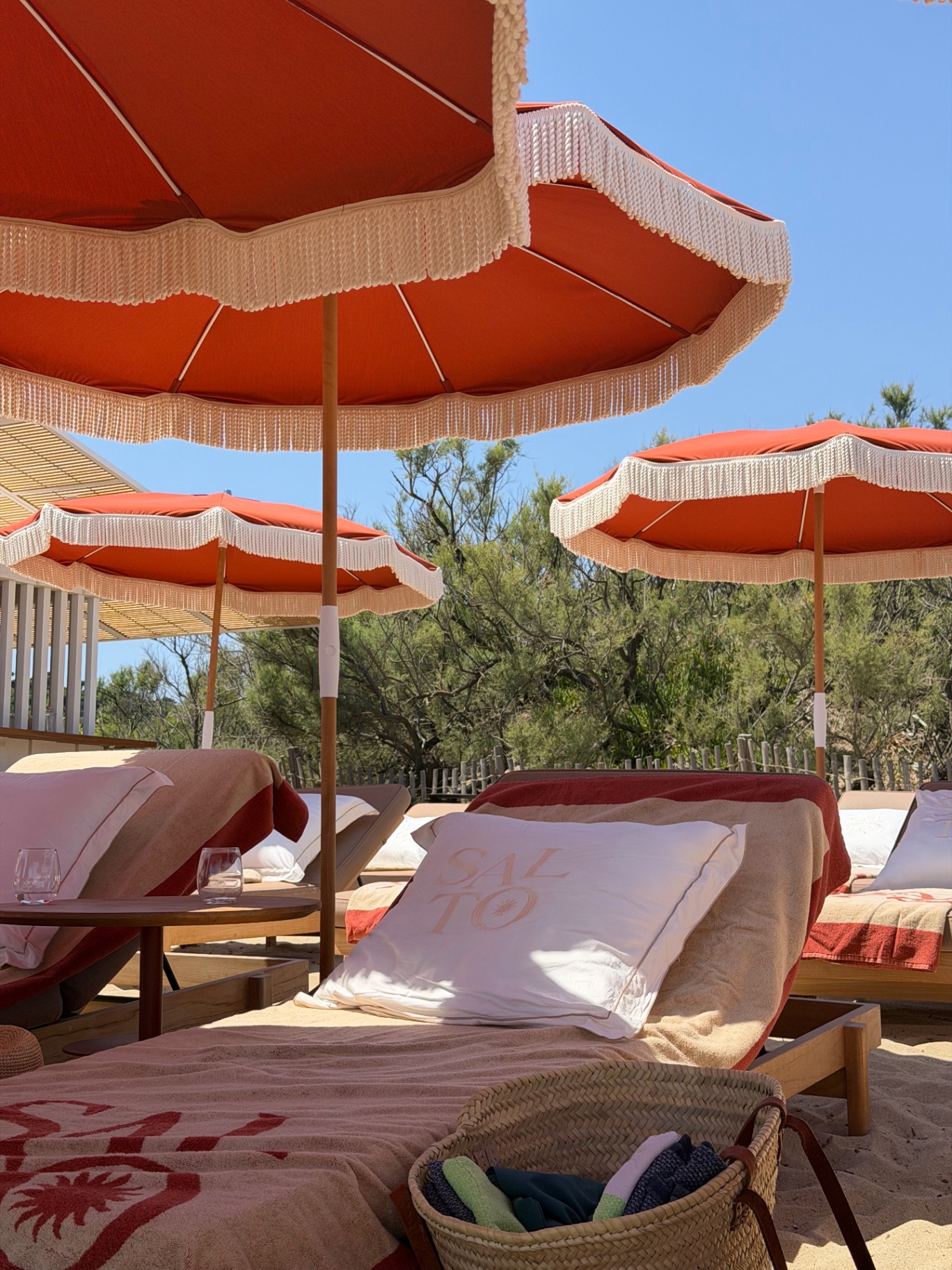 A SALTO-branded cushion on a double sun lounger under a terracotta fringed parasol at SALTO beach club, with a woven basket in the sand and trees behind.