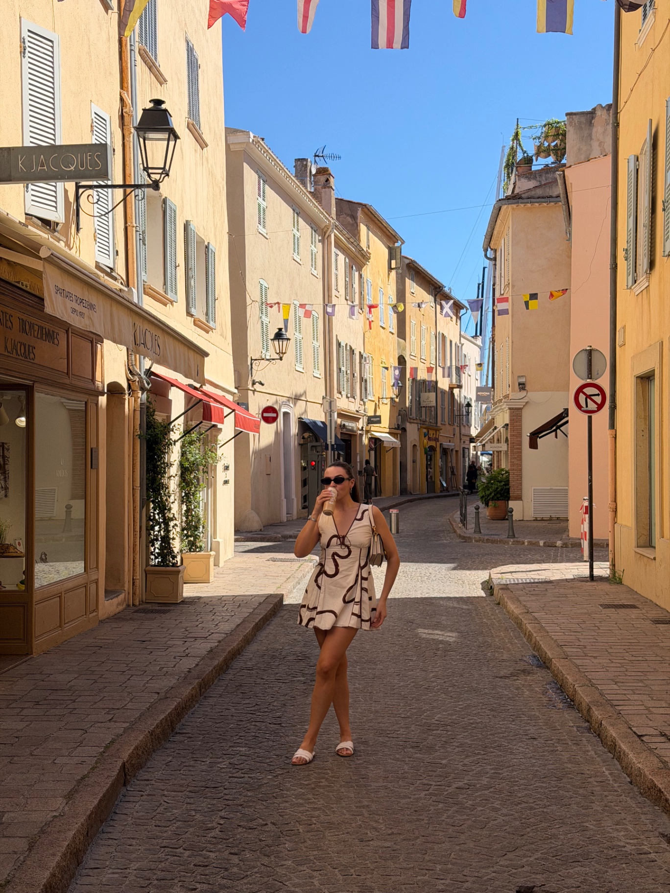 Holly wandering the sun-drenched cobblestone streets of Saint-Tropez Old Town, with pastel buildings and colourful pennant flags overhead.