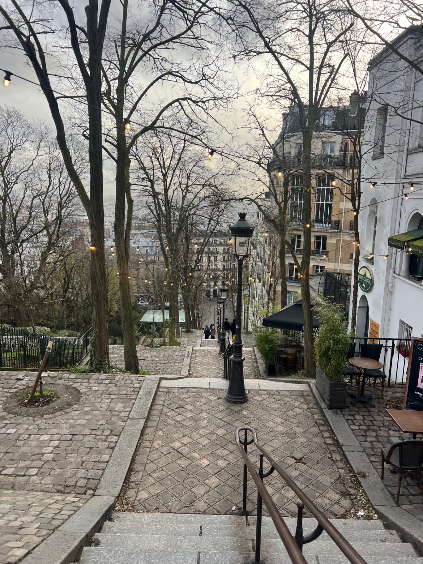 The Montmartre stepped street with vintage lamp posts, bare trees strung with festoon lights and cobblestones descending toward the city below.