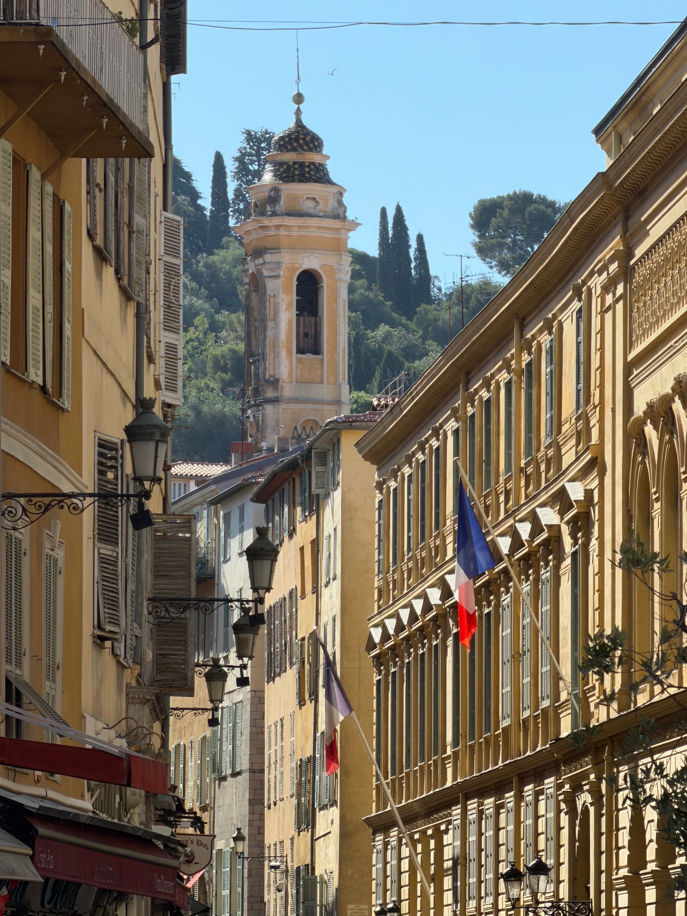 Sunlit ochre buildings and a baroque bell tower rising above a narrow street in Nice Old Town, French flag hanging from a façade.