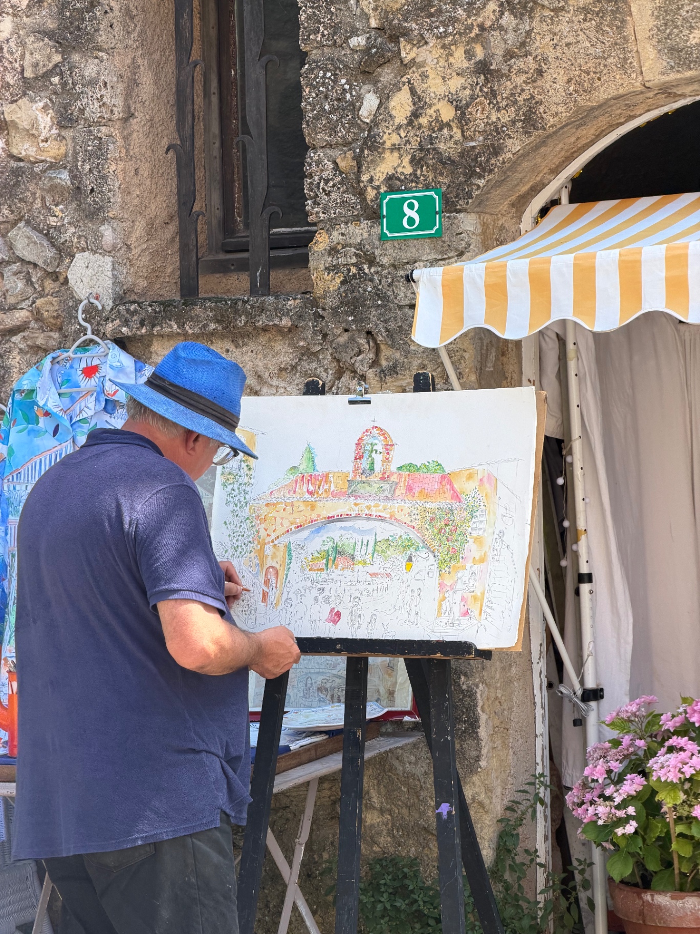 A local watercolour artist in a blue sun hat painting a scene of Tourtour's archway and bell tower on an easel, set up beneath a yellow-and-white striped awning outside a stone house numbered 8.